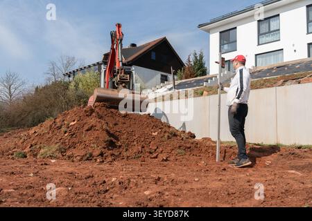 Uomo con tappo rosso che tiene l'asta di rilevamento accanto a un mini escavatore che scava terreno rosso in un cantiere residenziale. Lavori di livellamento e livellamento in Foto Stock