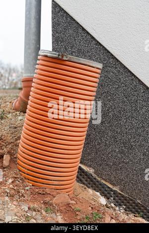 Un tubo di drenaggio spesso che sporge dal suolo vicino a una casa, parte del sistema di gestione delle acque, circondato da terreno ed elementi da costruzione. Alto Foto Stock