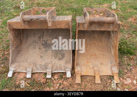 Primo piano di due benne usate posizionate sul terreno in un cantiere residenziale in pendenza. Un mini escavatore rosso e una casa sono visibili nella Foto Stock