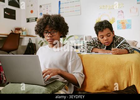 Adolescente di colore che studia su un notebook mentre il bambino scrive a casa Foto Stock