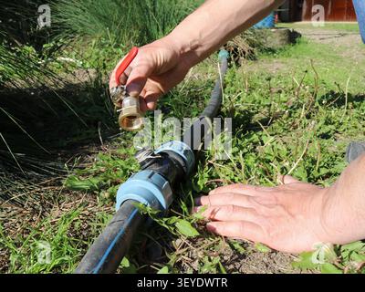 Le mani di un tecnico che collega una valvola a sfera in ottone con una maniglia rossa a un raccordo a T in plastica su un tubo in HDPE nero. Il sistema di irrigazione Foto Stock