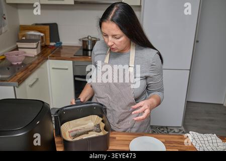 Una donna prepara il pesce fresco per la cottura posizionandolo in un cestello della friggitrice ad aria. Cucina casalinga sana e semplice in un ambiente moderno. Foto Stock