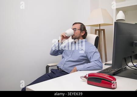 Un uomo professionale siede alla scrivania per bere un caffè durante una pausa in un moderno ambiente di lavoro. Foto Stock