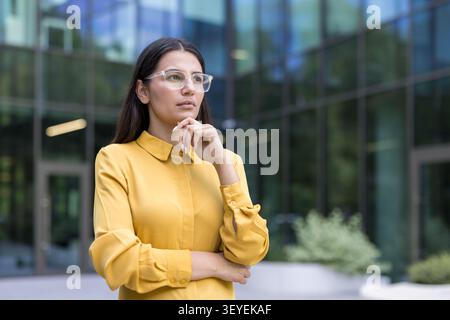 Donna d'affari professionista che indossa una camicia gialla e occhiali con montatura trasparente in piedi con una mano al mento, guardando lontano, riflettendo sulla strategia aziendale in un moderno edificio di uffici Foto Stock