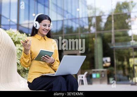 Una giovane donna che fa videoconferenze all'aperto con cuffie e laptop, discute di idee aziendali e prende appunti in uno stile di vita nomade digitale godendo di un lavoro flessibile Foto Stock
