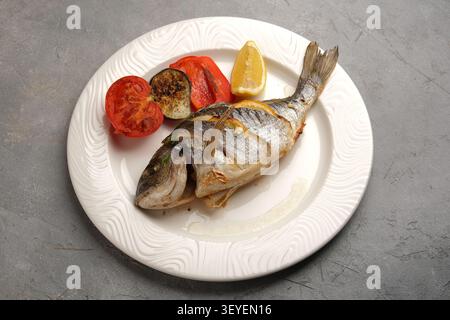 Pesce intero grigliato servito su piatto bianco con verdure arrostite e spicchi di limone Foto Stock