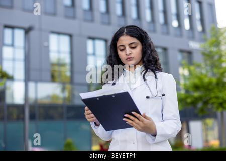 Giovane medico donna in un camice bianco e stetoscopio si concentra sulla revisione delle cartelle cliniche dei pazienti su un appunti, in piedi fuori da un moderno edificio clinico Foto Stock