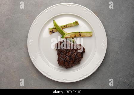 Bistecca alla griglia con zucchine carbonizzate su piastra in ceramica bianca su sfondo in pietra grigia Foto Stock