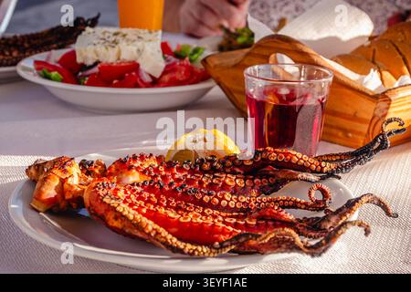 Polpo alla griglia servito con limone, accompagnato da bevande in un ambiente costiero durante l'estate. Foto Stock