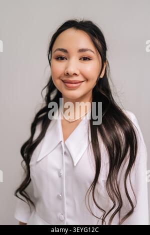 Allegra estetista asiatica in uniforme bianco, che rappresenta una cosmetologa o un medico sicuro di sé e professionale che offre bellezza e Foto Stock