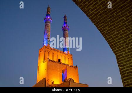 Una vista ad angolo basso parzialmente incorniciata da un arco di mattoni sulla destra, che mostra un'alta torre illuminata sormontata da due splendidi minareto a mosaico blu e viola Foto Stock