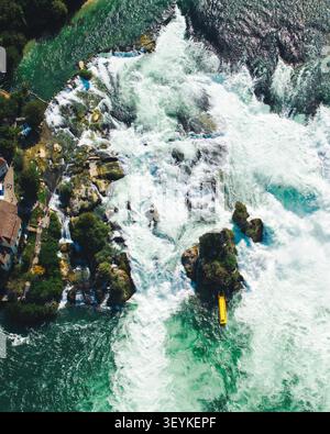 Vista aerea delle Cascate del Reno con potenti cascate d'acqua, affioramenti rocciosi, vegetazione lussureggiante e una piccola barca gialla che naviga sul fiume a Neuhausen Foto Stock