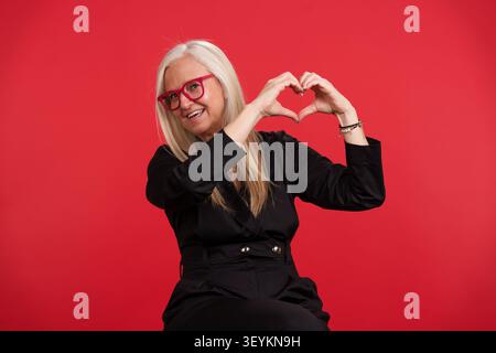 Donna matura che fa il simbolo del cuore esprimendo amore e cura Foto Stock