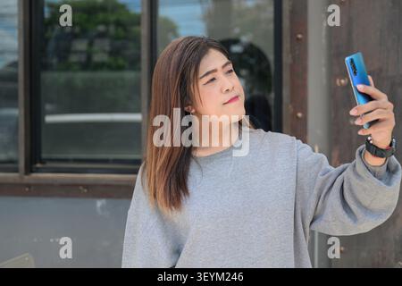 La donna transgender thailandese del sud-est asiatico nei suoi 40 anni documenta il suo stile di vita digitale mentre posa vicino a una struttura di pannelli di finestre. Vestito con una tuta grigia Foto Stock