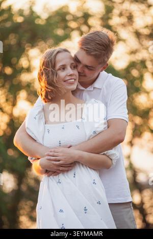 Un giovane uomo abbraccia dolcemente una donna sorridente da dietro, immersa nel soffice bagliore della luce del tramonto in mezzo a una vegetazione sfocata Foto Stock
