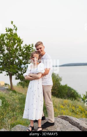 Una felice coppia adolescente si trova vicino su un pendio erboso con un grande specchio d'acqua sullo sfondo, godendosi un momento di pace all'aperto Foto Stock