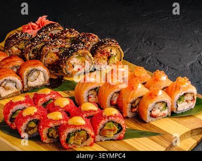 Un grande piatto mostra vari sushi roll disposti ordinatamente su un asse di legno. I rotoli includono riempimenti e guarnizioni diversi. Lo zenzero rosa si trova accanto al sushi. Foto Stock