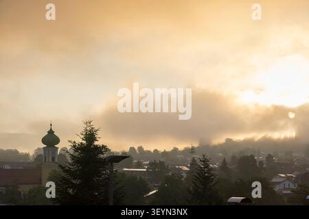 Nebbia mattutina illuminata dall'alba su una piccola cittadina bavarese con la torre della chiesa e le colline circostanti. Foto Stock