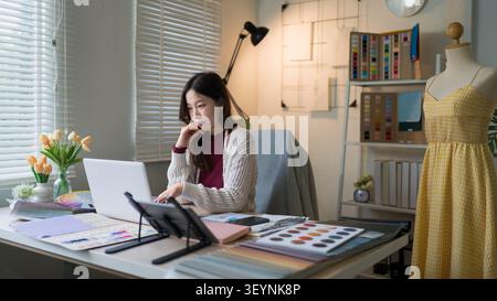 Stilista asiatico che lavora con campioni di colore e tessuti in uno studio luminoso e moderno con manichino e laptop Foto Stock