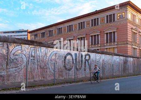 Berlino, Germania: il ciclista solitario passa davanti ai graffiti "Save Our Planet" su una sezione del muro iconico di Berlino, uno storico confine urbano d'epoca. Foto Stock