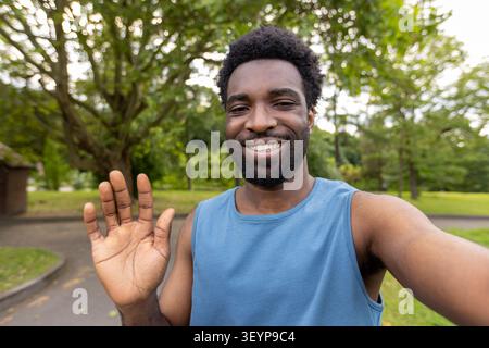 Giovane felice che si diverte all'aria aperta, effettua una videochiamata o saluta le persone tenendo in mano uno smartphone e sorridendo direttamente davanti alla fotocamera in un parco pubblico Foto Stock