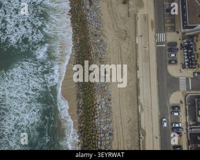 Veduta aerea di Praia do Furadouro che mostra la spiaggia sabbiosa, la diga di pietra, le onde che si infrangono e la strada costiera adiacente con auto parcheggiate a Ovar, Aveiro, Foto Stock