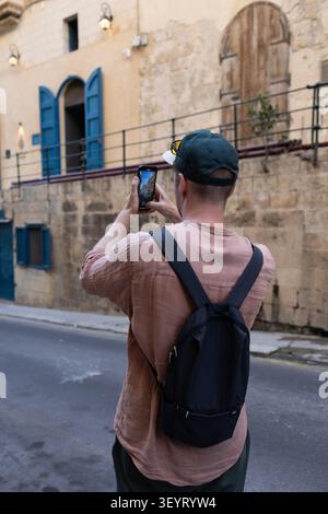 LA VALLETTA, MALTA-GIUGNO 2026: Turista con zaino che scatta foto di strade storiche utilizzando lo smartphone. Foto Stock
