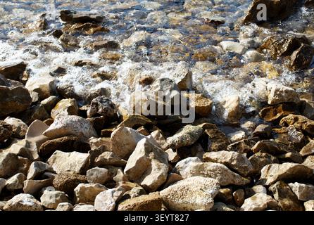 Primo piano di acqua di mare limpida che scorre su rocce naturali lungo una costa rocciosa mediterranea. La luce del sole evidenzia le pietre strutturate e le onde gentili Foto Stock