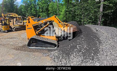 Una pala cingolata compatta gialla che sposta un mucchio di ghiaia su un cantiere in attività circondato da alberi. Foto Stock