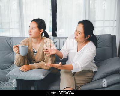 Donne asiatiche sedute sul divano a casa durante una conversazione seria, una tazza in mano mentre un'altra dà consigli, concetto di comunicazione familiare, sostegno, Foto Stock