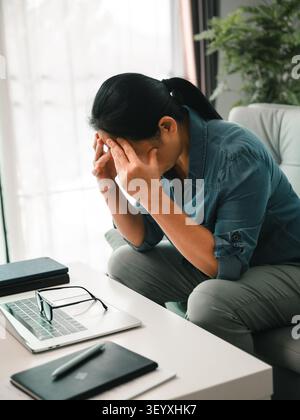 Donna asiatica stressata seduta sul divano con la testa in mano vicino a laptop, occhiali e notebook, che mostra esaurimento, stress finanziario, pressione di esercizio e ment Foto Stock