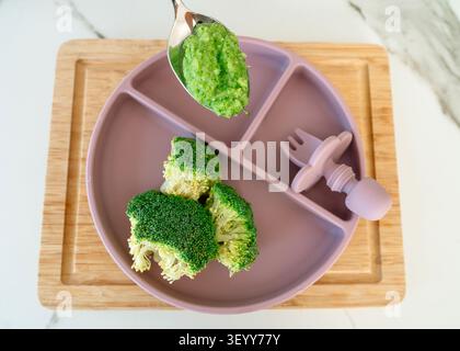 Una piastra per bambini in silicone viola con fiori di broccoli verdi freschi e un cucchiaio ripieno di purea di verdure. Posizionato su un asse di legno per lo svezzamento guidato dal bambino Foto Stock