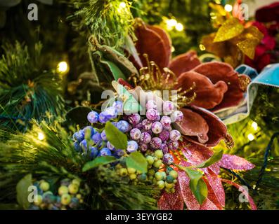 Primo piano di eleganti decorazioni sull'albero di Natale con frutti di bosco artificiali smerigliati viola e blu, ornamento di fiori di velluto e calde luci fiabesche Foto Stock