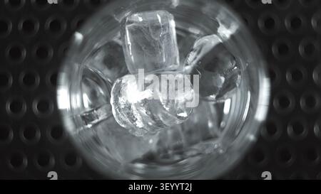 Cubetti di ghiaccio che si sciolgono in un bicchiere trasparente, mostrando liquido freddo rinfrescante e acqua ghiacciata con uno sfondo scuro astratto Foto Stock