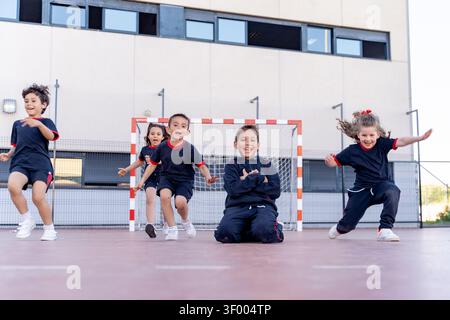 I bambini in uniforme saltano e ridono in un parco giochi all'aperto della scuola, esprimendo gioia ed energia durante una partita Foto Stock