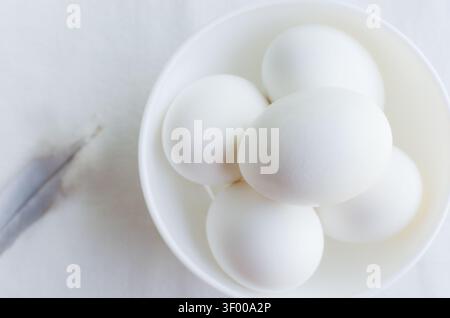 Composizione minimalista con una ciotola bianca riempita con cinque uova bianche, una piuma leggera accanto ad essa. Puro stile monocromatico, enfasi sulla semplicità, Foto Stock