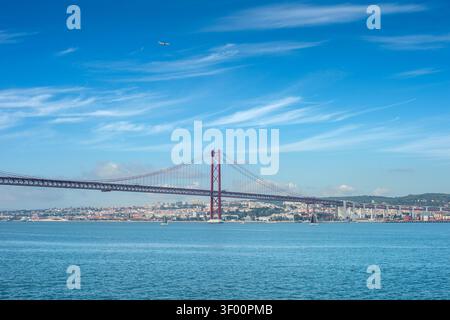 Un grande ponte sospeso rosso, il 25 aprile, si estende attraverso un ampio fiume con navi in acqua e uno skyline della città Foto Stock