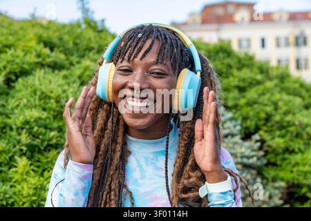 Ritratto ravvicinato di una giovane donna sorridente con capelli intrecciati che indossa cuffie sovrauricolari colorate all'aperto, tenendo i padiglioni mentre ascolti i mu Foto Stock
