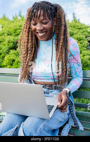Giovane donna allegra con lunghe trecce sorride mentre lavora su un notebook all'aperto, seduta su una panchina del parco in natura, indossando un top casual e colorato Foto Stock
