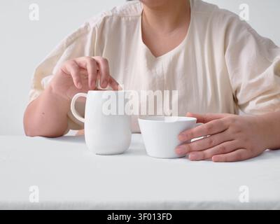 Vista ritagliata di una donna seduta su un tavolo bianco, che tiene una tazza in ceramica minimalista accanto a una piccola caraffa del latte. Luce naturale soffusa e tonalità neutre Foto Stock