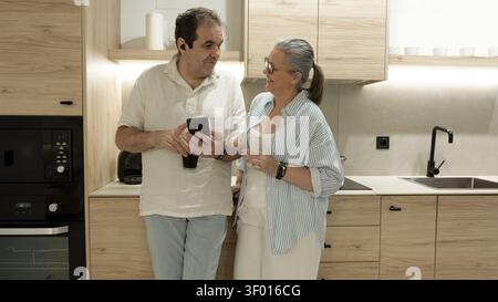 Una coppia matura si rilassa in una cucina moderna e luminosa, sorridendo e parlando mentre condivide uno smartphone e un caffè, mostrando la connessione quotidiana, il comfort e. Foto Stock
