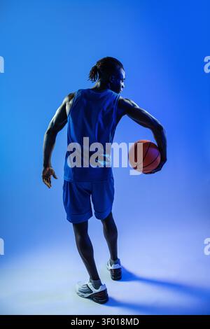 Vista posteriore completa di un giocatore di basket nero che tiene in mano una palla arancione, indossa una maglia blu e pantaloncini corti in uno studio blu con luci e luci spettacolari Foto Stock
