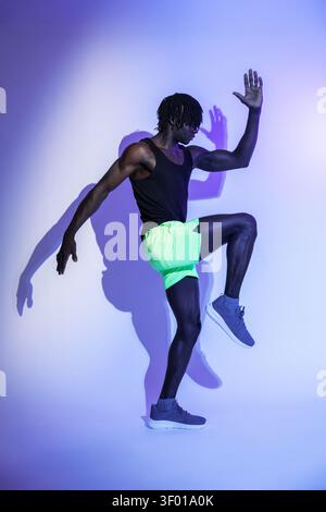 Atleta maschile di colore in pantaloncini al neon che esegue un riscaldamento elevato del ginocchio in uno studio, illuminato da una brillante luce blu e viola, che mostra forza e equilibrio Foto Stock