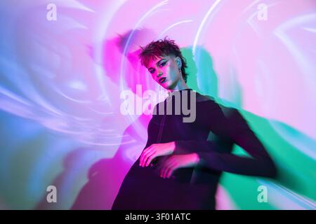 Elegante giovane donna cyberpunk che indossa un abito nero sotto una vivace luce rosa neon e color acqua, con sfocatura in movimento e bagliore olografico che creano un futuro Foto Stock