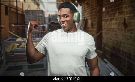L'uomo nero che indossa le cuffie si flette e sorride in strada vicino a ponteggi e pallet; musica serenità. Foto Stock