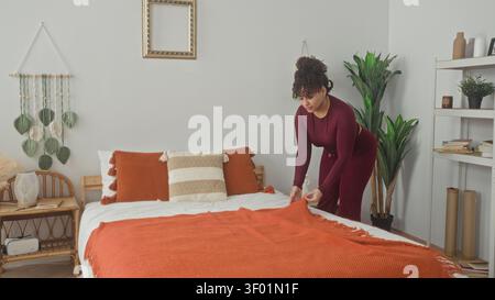 Donna che nasconde una coperta arancione sul letto con le mani e i cuscini liscianti mentre si piega vicino allo scaffale in una camera da letto dell'edificio; calma routine. Foto Stock