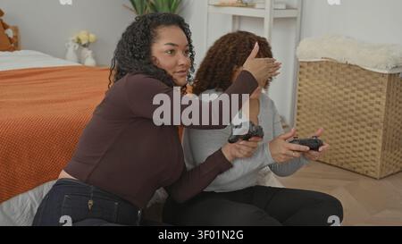 Due donne sul pavimento di una camera da letto che reggono controller di gioco, una mano copre il volto dell'altra mentre si avvicinano a un joystick e ridono; giocosa competizione Foto Stock