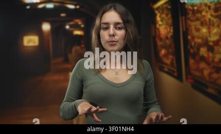 Donna con palme in alto e occhi chiusi nella galleria d'arte dell'edificio, indossa camicia verde e collana a cuore; riflesso calmo. Foto Stock