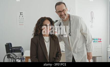 Il medico posiziona la mano sulla spalla del paziente mostrando lo stetoscopio come donna con gli occhiali sorride nella costruzione della clinica; cura rassicurante. Foto Stock