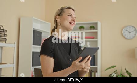 Donna che tiene il tablet con entrambe le mani, sorride e fa una pompa a pugno in studio con scaffale, orologio e pianta d'casa visibili; felicità celebrazione. Foto Stock